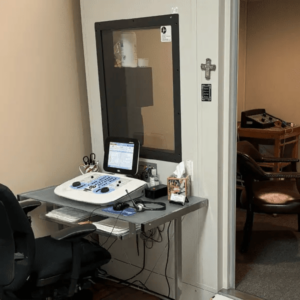 An audiologist's workstation featuring a hearing test audiometer with a screen, control panel, and headphones, positioned next to a soundproof booth with a window for patient observation